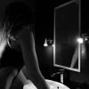 A woman leans over a sink in a dimly lit bathroom, reflecting in the mirror above.