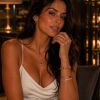 A woman sits confidently in an elegant setting, wearing a white dress and striking jewelry. Soft lighting enhances her features.