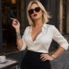 Una donna bionda in camicia bianca e gonna nera fuma un sigaretta, seduta a un tavolo con caffè.
