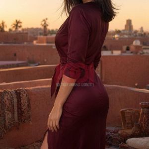 A woman in a burgundy dress stands on a rooftop at sunset, with a cityscape and palm trees in the background.