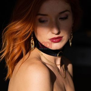Ritratto di una giovane donna dai lunghi capelli rossi, con orecchini dorati e un collare elegante, immersa in luce soffusa.