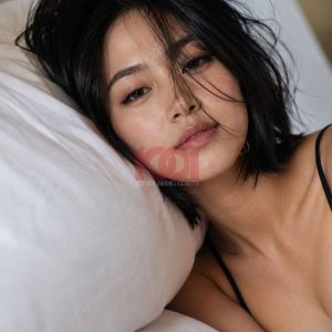 A woman with tousled hair rests on white pillows, gazing softly with a serene expression.