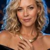 A woman with curly hair wears a black dress and gold necklace, softly smiling with a glimmering background.