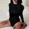 Giovane donna vestita di nero, con tatuaggi su cosce, seduta su un letto bianco, sguardo intenso e trucco scuro.
