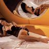 Immagine di una donna distesa su un letto, indossando un abito nero trasparente, con un grande specchio.